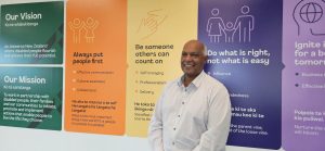 Angelo in white business shirt smiling infront of our vision, mission and values wall.