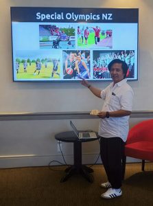 Andy is wearing a white top and dark pants, smilling while presenting his inspirational speech in front of an audience.