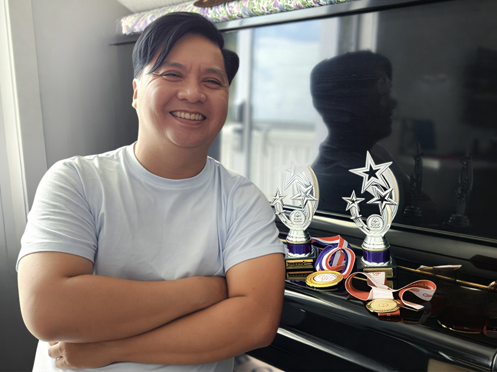 Andy is seated beside his piano, arms crossed, smiling. He is wearing a light blue shirt. His Special Olympics awards on display beside him.