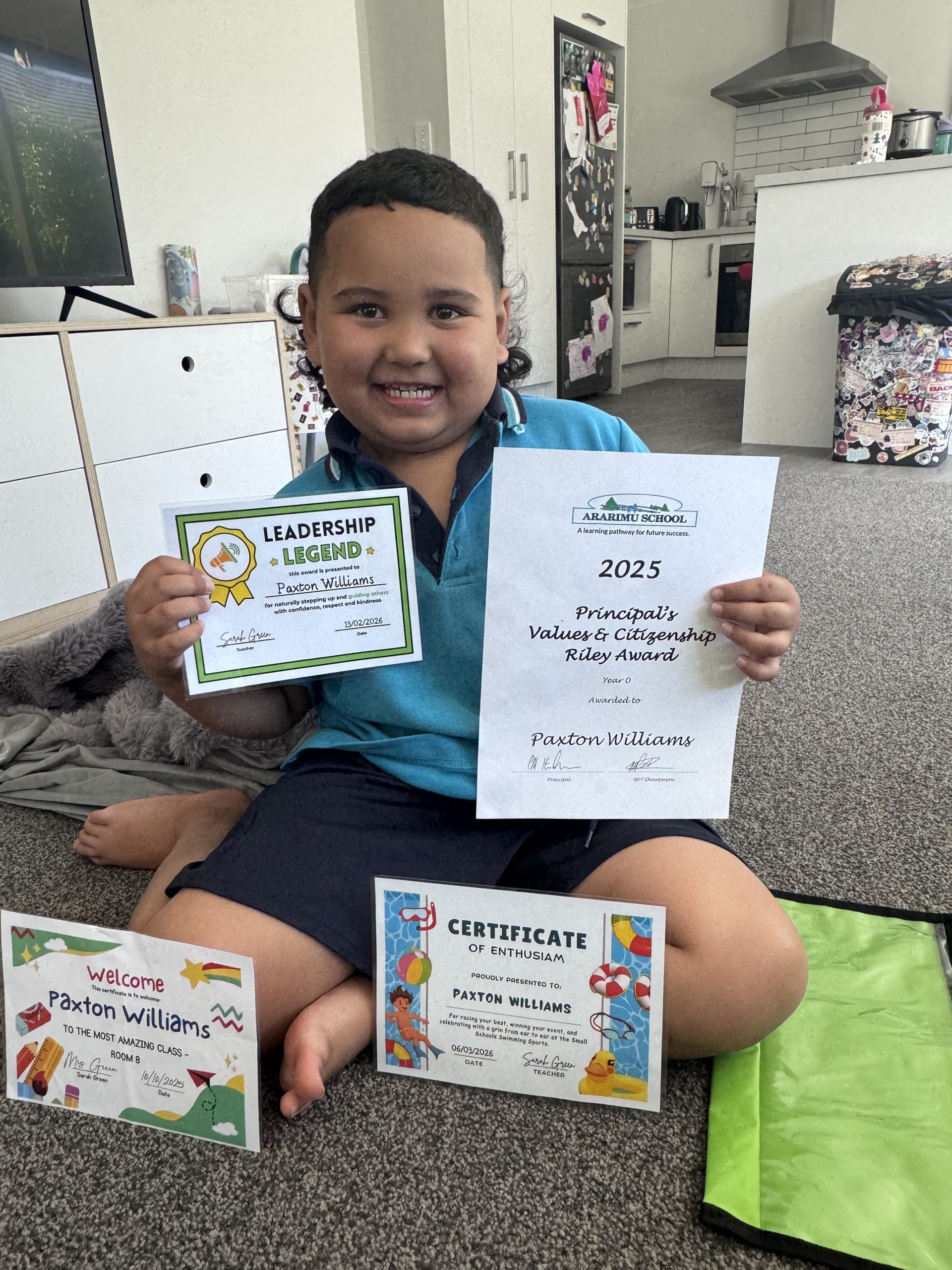 Paxton is seated on the floor, smiling and proudly showing 4 leadership awards from school