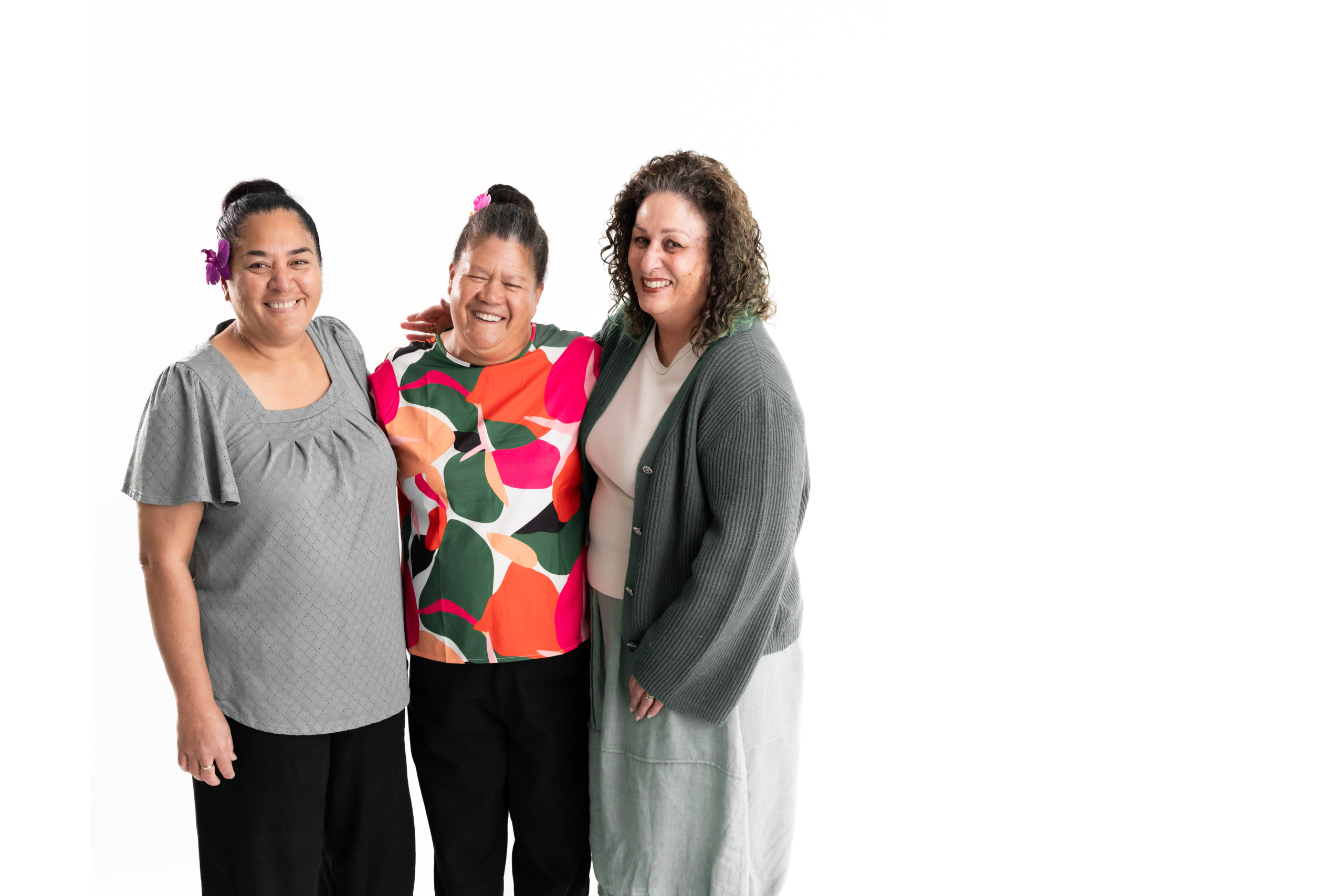 Three of our female employees standing closely together and smiling happily at the camera. They have Pacific and Maori ancestry, and are wearing shades of green.